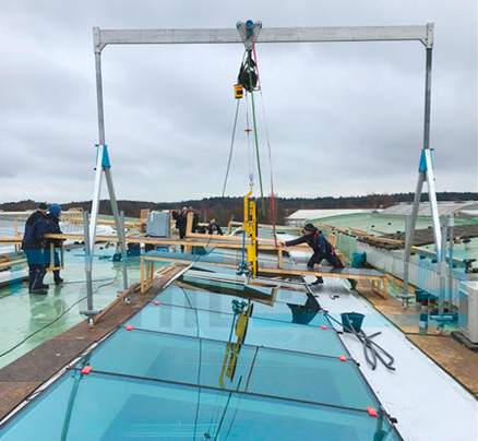 Auf einer Baustelle installieren Arbeiter mit einem SCHILLING Alu-Portalkran große Dachfenster auf dem Dach eines Firmengebäudes.