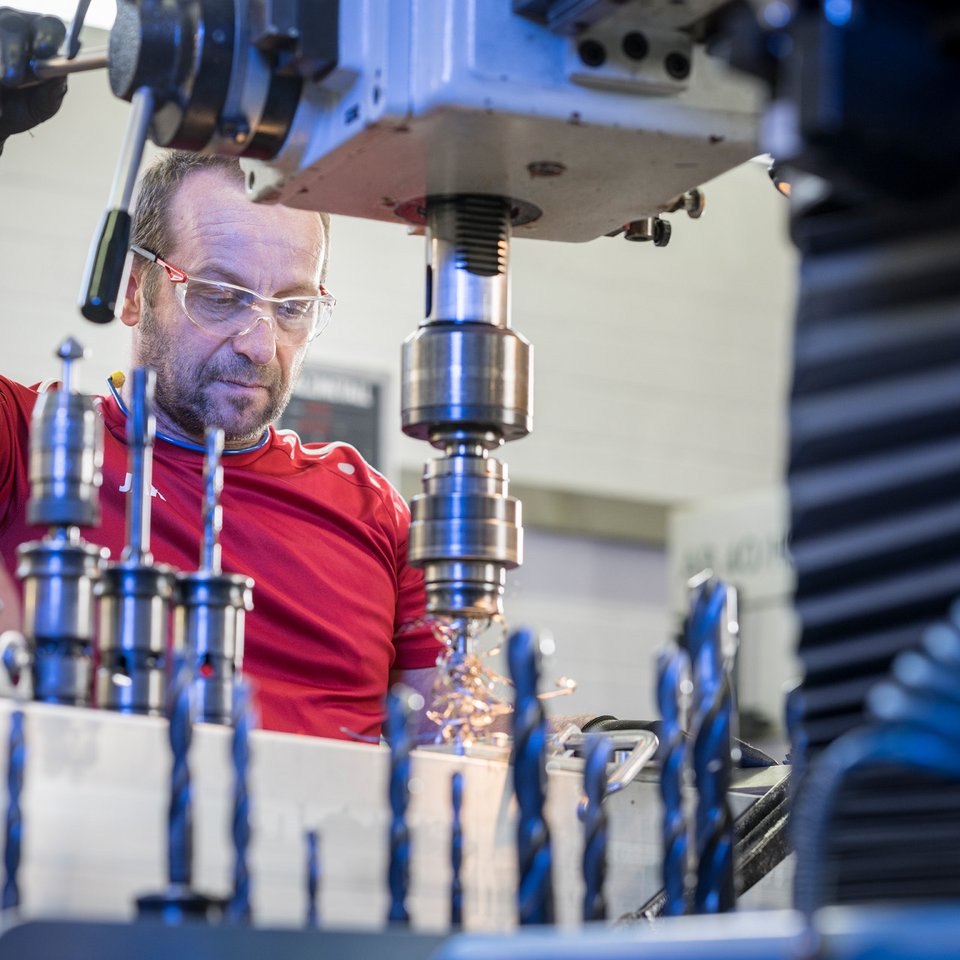Ein Mann bedient einen Bohrer, der Aluminium bearbeitet, Späne fliegen, in Vordergrund mehre Bohrköpfe.