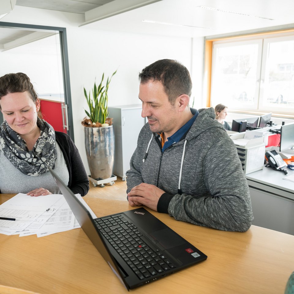 Zwei Personen besprechen gemeinsam Papiere und einen Laptop auf einem Tisch in einem hellen Büro, im Hintergrund arbeiten weitere Personen an Schreibtischen.