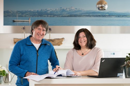 Martin und Heike Schilling stehen nebeneinander, lächeln und schauen in die Kamera, vor ihnen geöffnete Unterlagen, daneben steht ein Laptop.