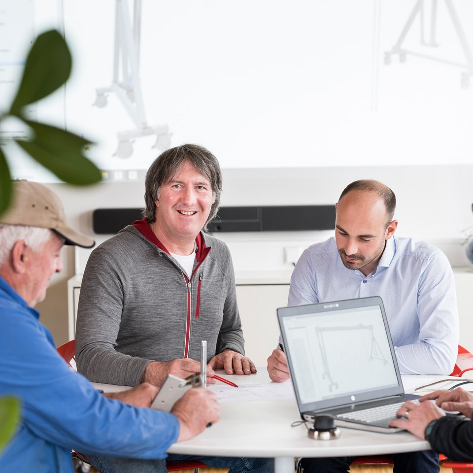 Martin Schilling sitzt gemeinsam mit Mitarbeitern an einem Tisch und arbeitet gemeinsam an technischen Skizzen, eine Person nutzt einen Laptop.