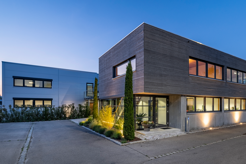 Der moderne, zweistöckige Firmensitz von SCHILLING Kran- und Hebetechnik in Friedrichshafen mit großflächigen Fenstern und Holzverkleidung, beleuchtet in der Abenddämmerung, umgeben von Straße und Gehweg.