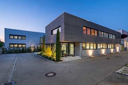 Der moderne, zweistöckige Firmensitz von SCHILLING Kran- und Hebetechnik in Friedrichshafen mit großflächigen Fenstern und Holzverkleidung, beleuchtet in der Abenddämmerung, umgeben von Straße und Gehweg.
