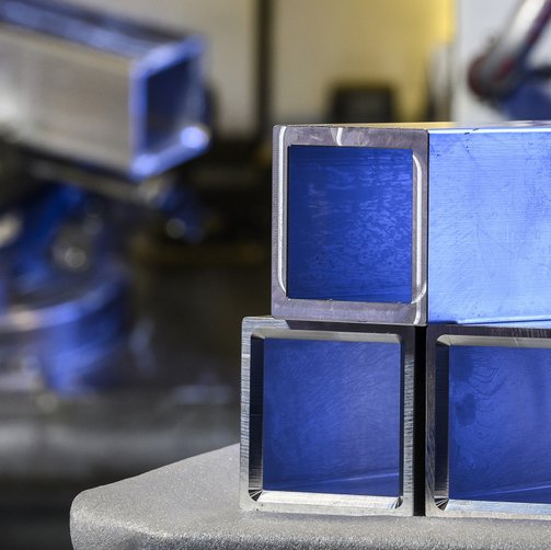 Vier ineinander gesteckte, glänzende quadratische Rohlinge aus Aluminium liegen auf einer Fläche, im Hintergrund unscharf Maschinenbauteile.